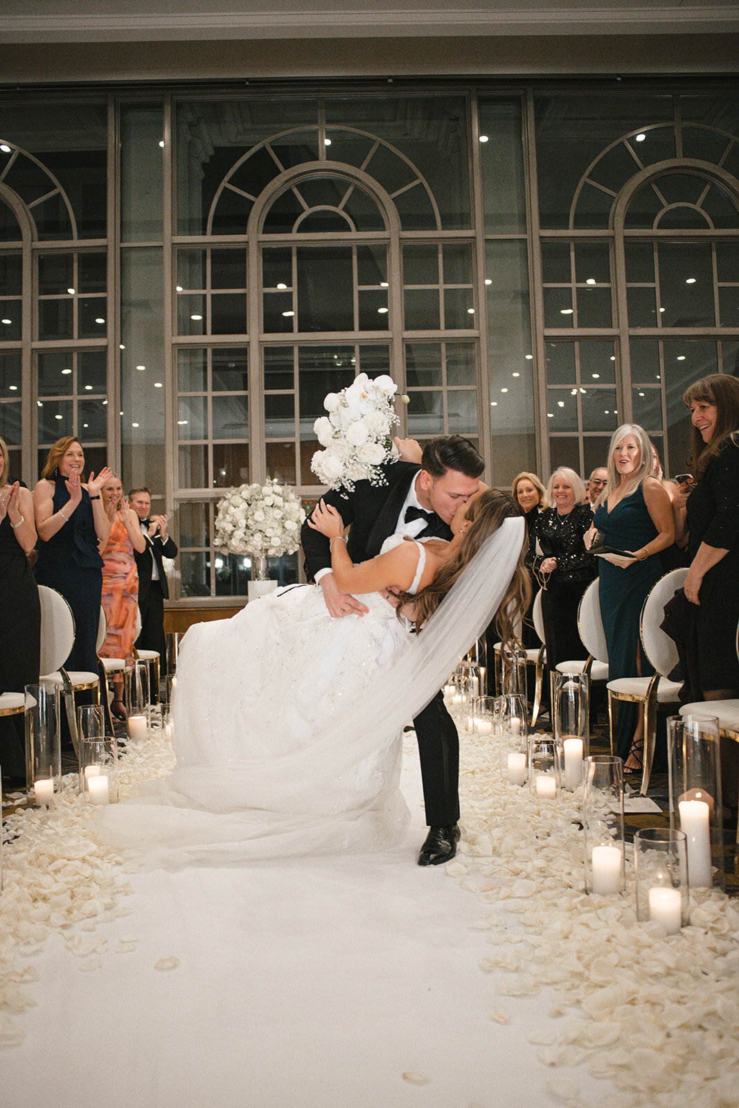 Ballroom first-kiss moment along a white rose-petal aisle